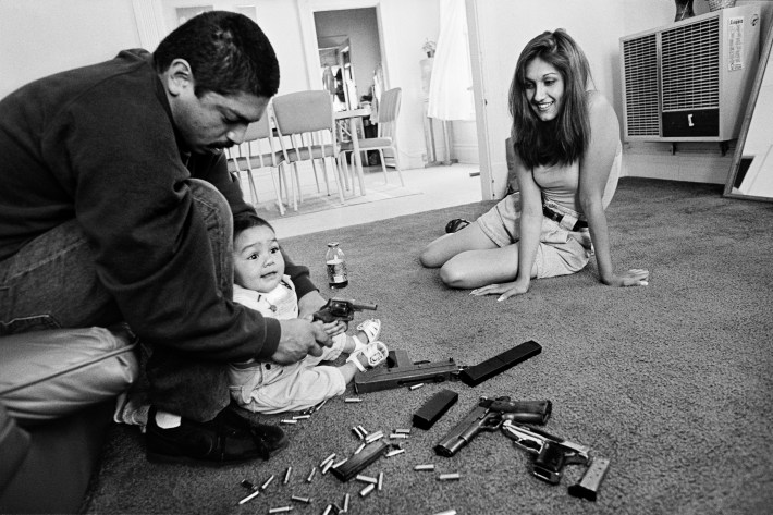 The morning after a rival gang tried to shoot Chivo for the fourth time. Chivo teaches his daughter how to hold a .32-caliber pistol. Her mother looks on. Boyle Heights, Los Angeles, CA, 1993