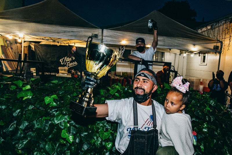 Vic Villa holds his daughter while he raises the 2021 trophy and someone behind him raises a can of model.