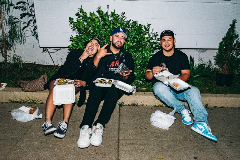 Three people with styrofoam containers on their lap enjoying tacos while posing for the camera.